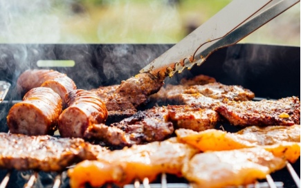 Meat is sizzling on a grill, with sausages and steaks cooking, while tongs turn a piece. Smoke rises, suggesting an outdoor barbecue setting with blurred greenery in the background.