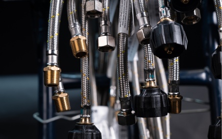 Braided metal hoses hang vertically, featuring various brass and black plastic fittings. They are suspended in an industrial setting with a dark, blurred background, suggesting utility or equipment assembly.