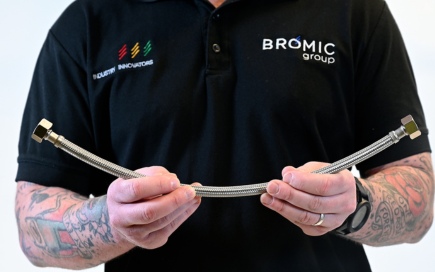 A person holds a flexible metal hose, bending it slightly. They wear a black shirt with logos reading 