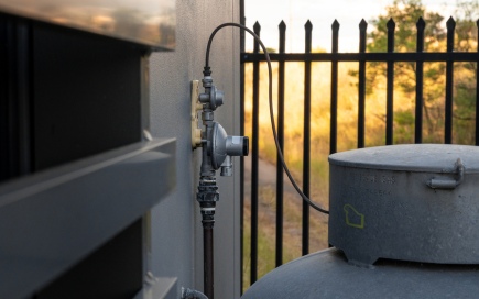 A metal regulator is attached to a wall, connected by cables to a nearby cylindrical tank. The background shows a black iron fence and sunlit grassy field.