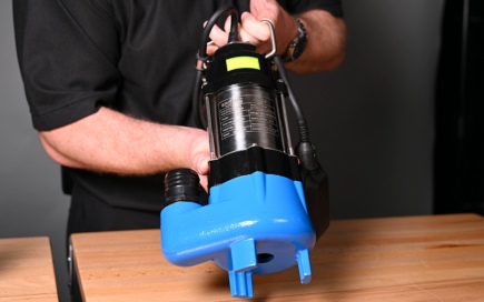 A person holds a blue and black submersible pump above a wooden table, demonstrating the equipment against a plain, dark background.