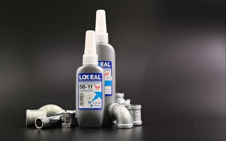 Two bottles of Loxeal engineering adhesives stand upright among scattered metal pipe fittings on a dark surface. The visible label reads: 