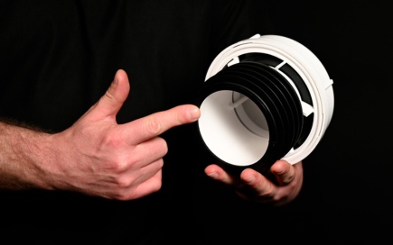 A person holds and points at a cylindrical black and white plastic object, possibly a component or device part. The background is dark, highlighting the object and hands.