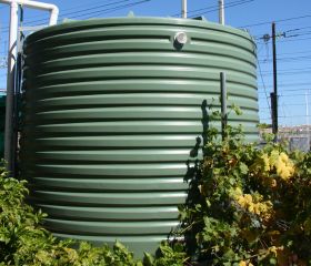 A large, green corrugated water tank stands stationary, surrounded by lush green and yellow foliage, with utility poles and wires visible in the background under a clear blue sky.
