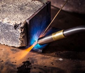 A welding torch emits a blue and orange flame onto a metal cube, while resting on a workbench. The action creates intense heat for metalwork in an industrial setting.