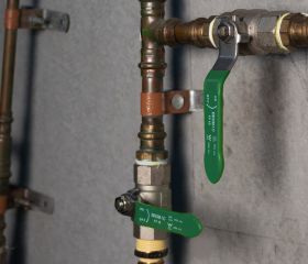 Pipes with green lever valves are mounted on a wall. The valves are labeled: 