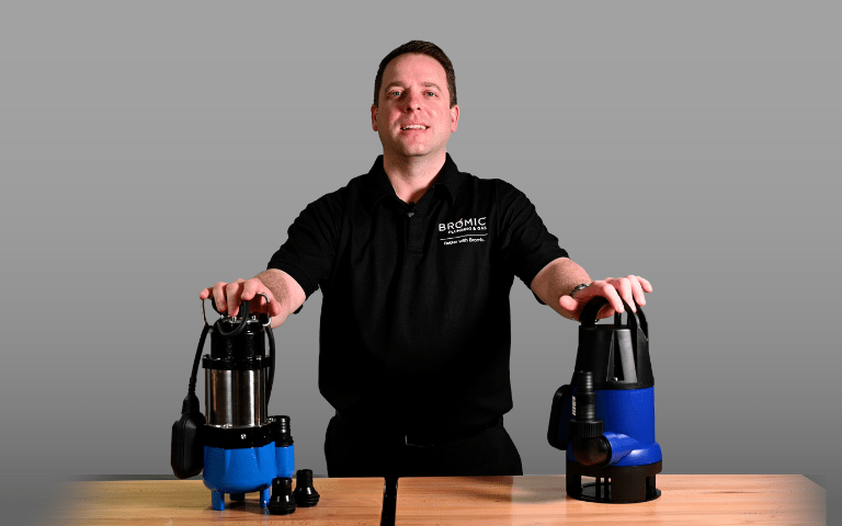 A person stands behind two blue sump pumps on a wooden table, demonstrating the products. The shirt reads, 