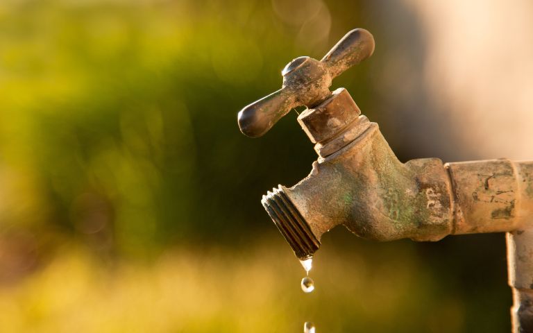 A rusted outdoor water faucet slowly drips with water in a sunlit garden setting, surrounded by a soft-focus background of green foliage.